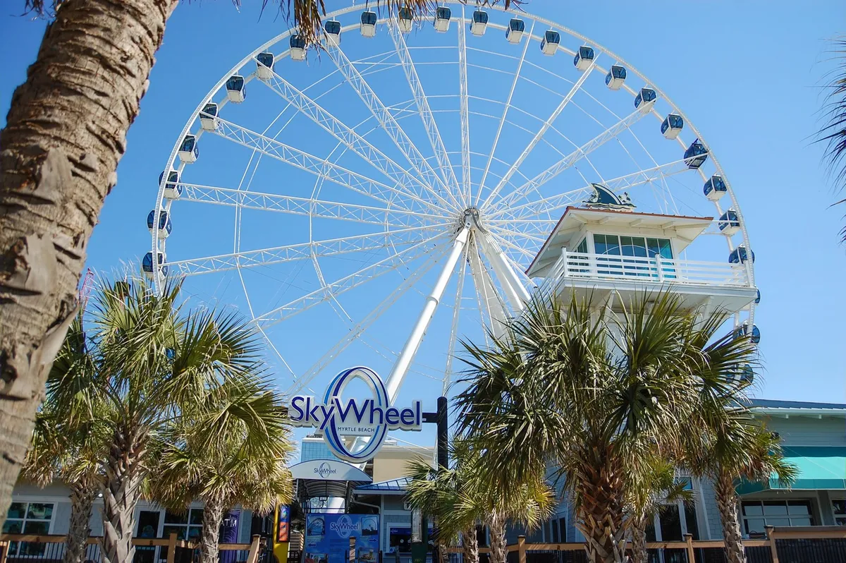 A vibrant exterior shot featuring Myrtle Beach’s iconic SkyWheel, ticket booth, and surrounding palm trees. Visitors can be seen excitedly preparing for a ride, with the blue sky and ocean nearby, capturing the thrill of local attractions.