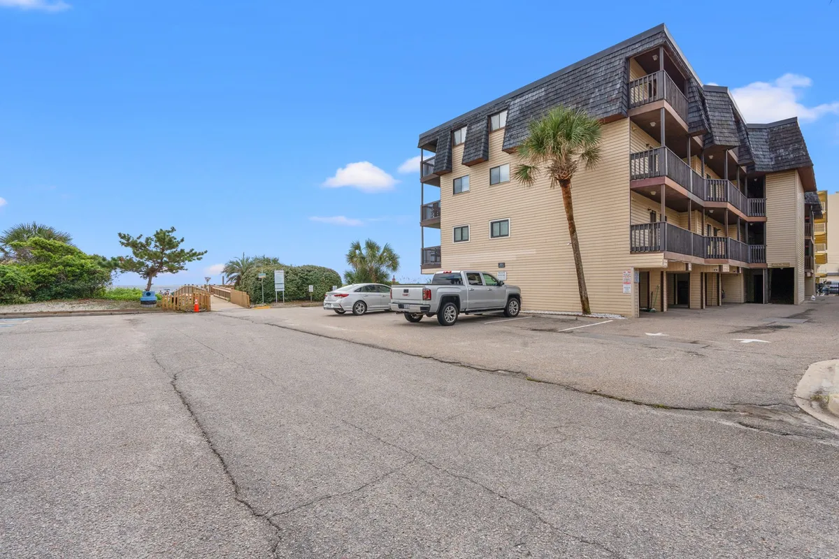 This exterior image shows the apartment building surrounded by palm trees, with a visible parking area in the foreground. The setting is casual, with the building's balconies suggesting private spaces to relax and enjoy the outdoors. The mood is welcoming and laid-back, reflecting a comfortable coastal lifestyle.