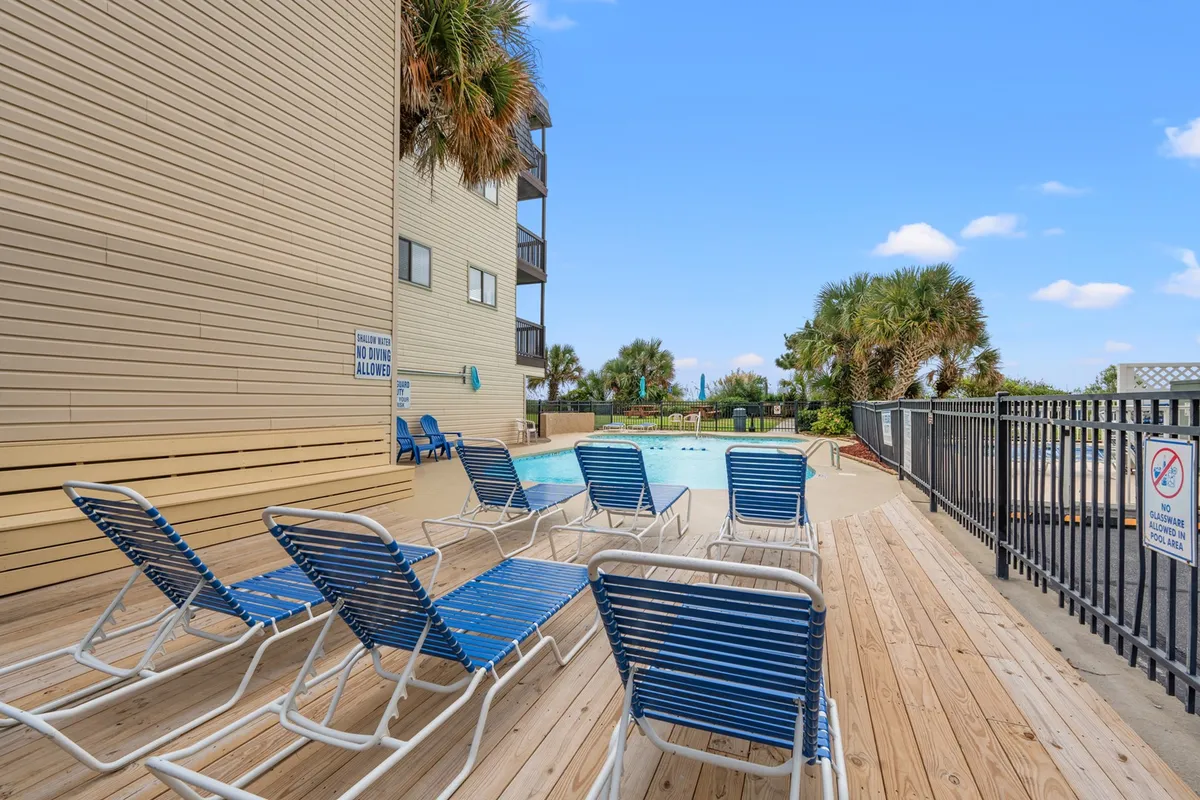 The inviting outdoor swimming pool of Long Bay Dunes lined with lounge chairs and flanked by palm trees. The pool area is calm yet vibrant, exuding a family-friendly vacation vibe, with the condo building in the background and plenty of space for sunbathing or playing.