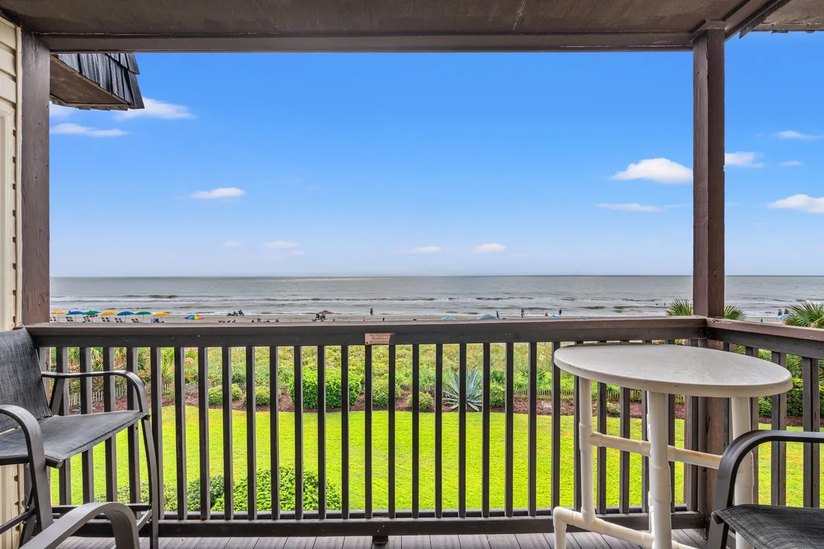 A private balcony is in the foreground, with its railing framing a spectacular view of the Atlantic Ocean, lush gardens below, and beachgoers enjoying the shore. The scene captures the peacefulness of the ocean breeze and the serenity offered by the balcony space connected to the main living area.