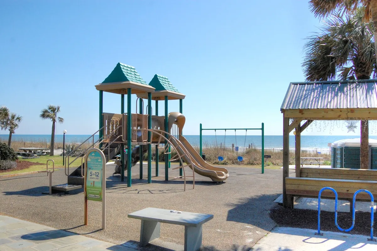 A sunny, vibrant playground area overlooking the ocean, complete with swings, picnic tables, and children playing. The scene radiates family-friendly energy and showcases both the coastal landscape and spaces for memorable outings.