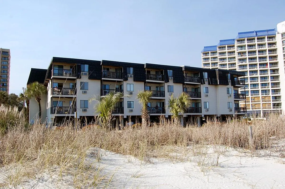 A beachfront building framed by palm trees and soft sand dunes, with balconies offering direct ocean views. The nearby high-rise hints at the area's lively resort atmosphere. The mood is relaxed and vacation-friendly, perfectly capturing the essence of oceanfront living.