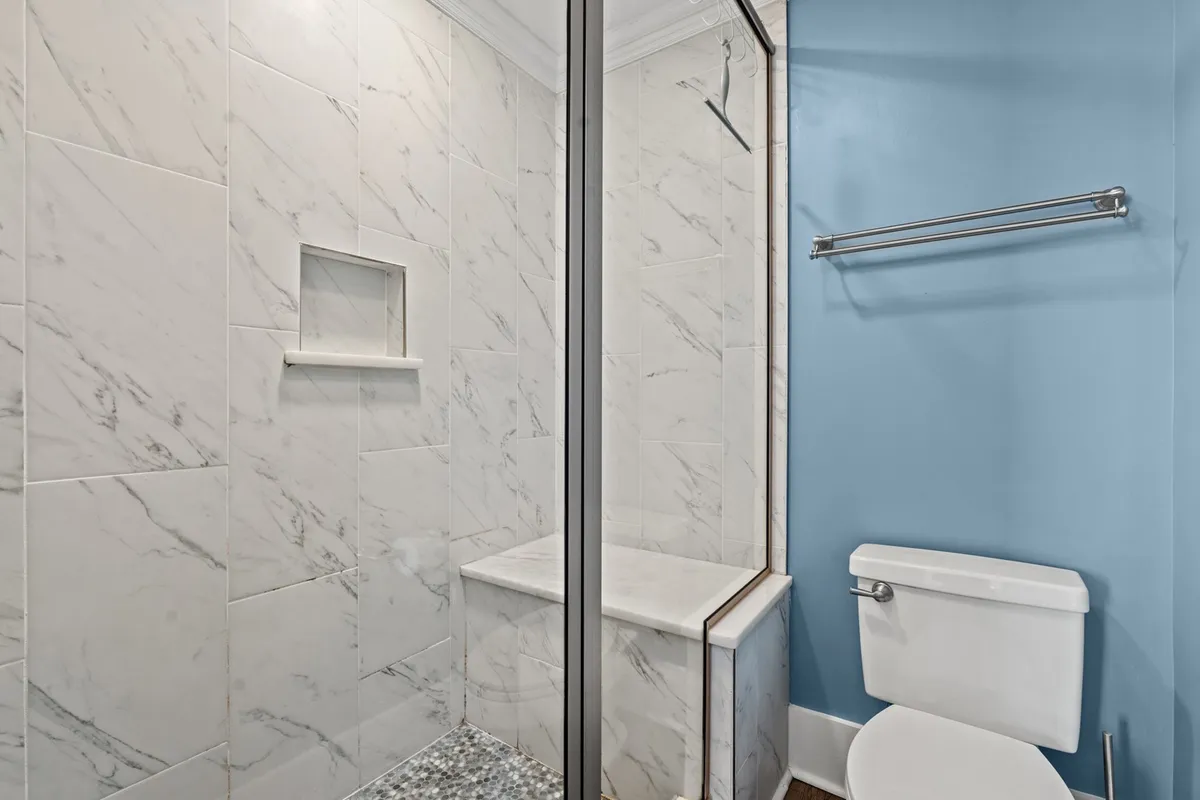 A bright, modern bathroom with a glass shower enclosure, marble tile walls, built-in shelf, white toilet, and a blue accent wall. The space feels fresh and inviting, perfect for unwinding after a long day.