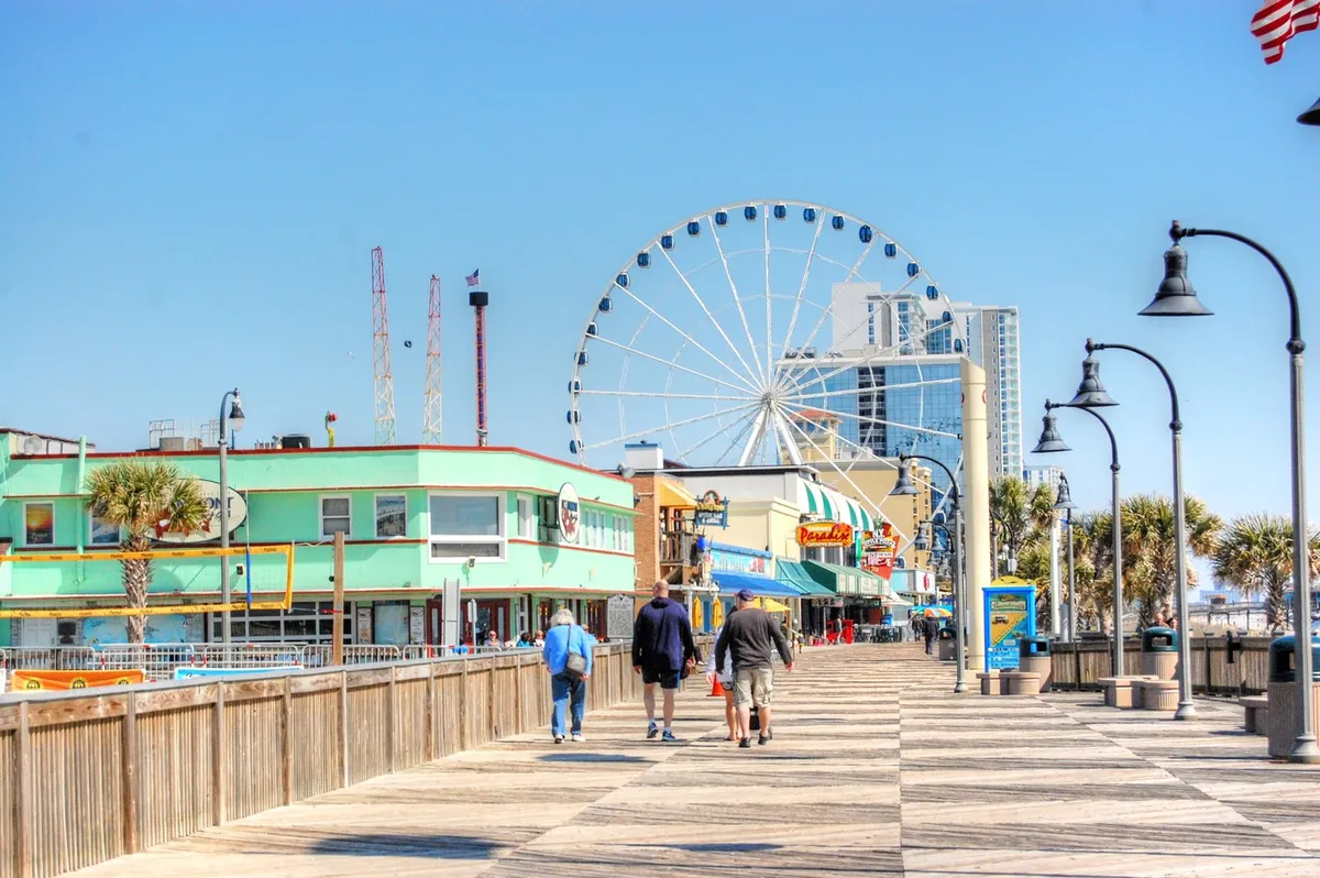 A vibrant boardwalk scene bustling with people, colorful buildings, and a towering Ferris wheel, evoking the energy of Myrtle Beach's nightlife, shopping, and entertainment options. The lively mood highlights the social, festive side of the area.