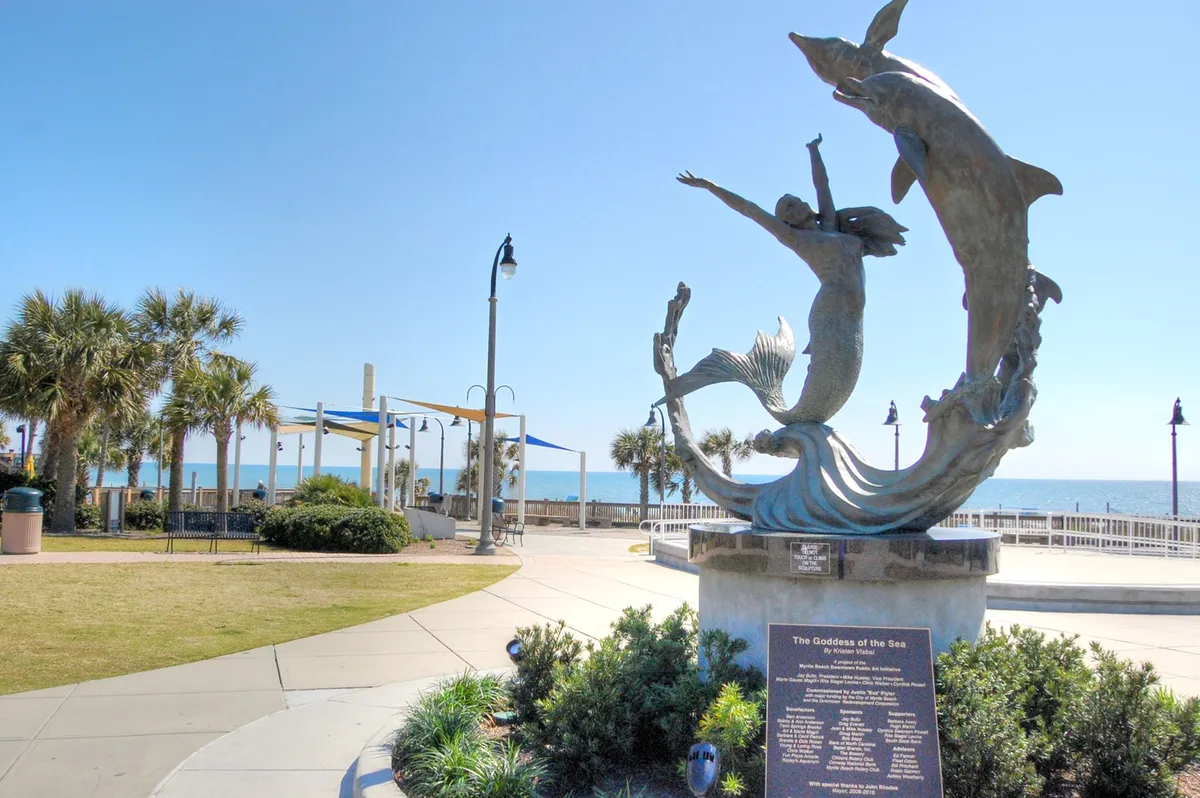 This image features the iconic Goddess of the Sea sculpture positioned along the Myrtle Beach Boardwalk, with palm trees, the ocean, and the famous boardwalk in view. The scene captures people strolling, taking photos, and relaxing near the waterfront, with the lively atmosphere of a popular local attraction.