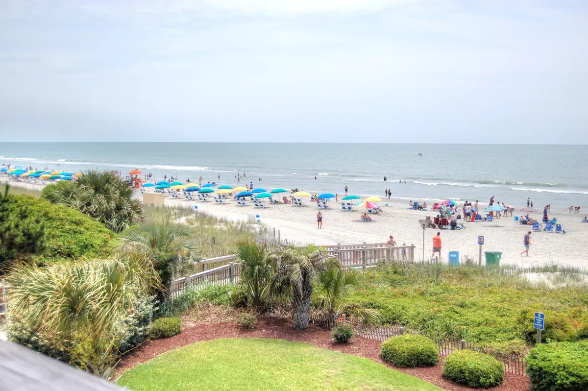 A bright, lively scene of families and friends enjoying Myrtle Beach. The image is filled with colorful beach umbrellas, people playing beach games, kids building sandcastles, and groups swimming in the ocean. Rolling sand dunes and gentle ocean waves set a classic beach day backdrop.