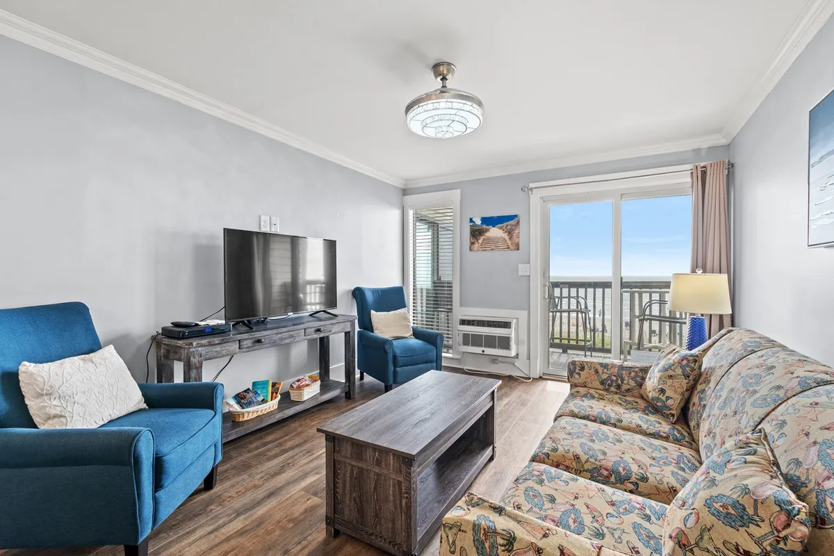 This image focuses on a cozy sofa facing a television, with a coffee table set up for activities like board games or snacks, and sunlight streaming through the balcony doors that offer a glimpse of the ocean view outside. The ambiance is warm and inviting, ideal for relaxing, watching movies, or socializing with friends and family.