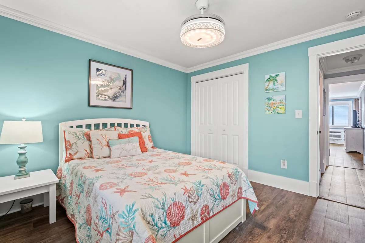A bedroom with a plush queen bed adorned in coral-patterned bedding, set against a calming teal accent wall. Decorative lamps and soothing artwork add to the tranquil atmosphere, and natural light softly fills the space.
