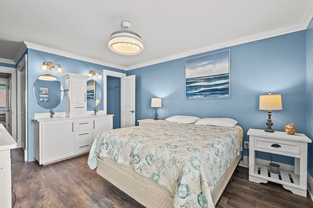 A second bedroom featuring a double bed with a patterned coverlet, blue painted walls, ocean-themed artwork, and a vanity with mirrors and lights. The space is filled with natural light, and the decor gives a refreshing, beachy vibe.